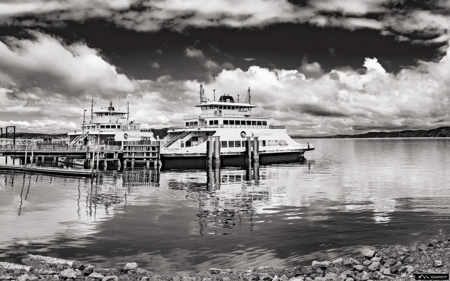 Two Steilacoom ferries are docked waiting for passengers for Anderson Island. The sky is full of clouds and the rocky shore is at the foreground.