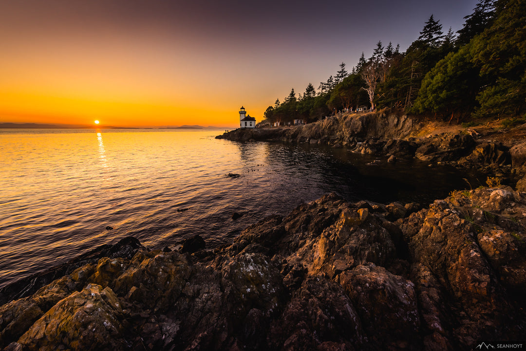 A warm sunset over rippling water with a lighthouse at center and large boulder rocks in foreground